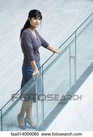 Stock Image - Woman standing with hands on glass guard rail, full length portrait. Fotosearch