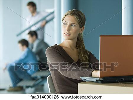 Stock Photograph - Woman using laptop on cardboard box, colleagues in background. Fotosearch