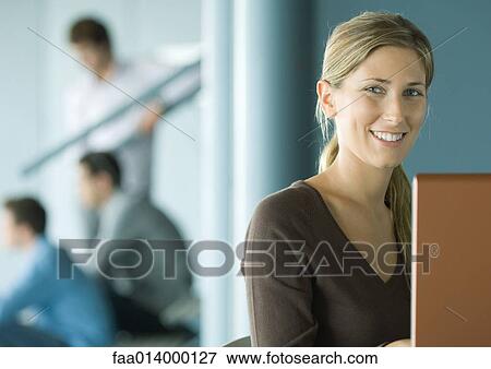 Stock Photo - Woman using laptop, smiling, colleagues in background. Fotosearch