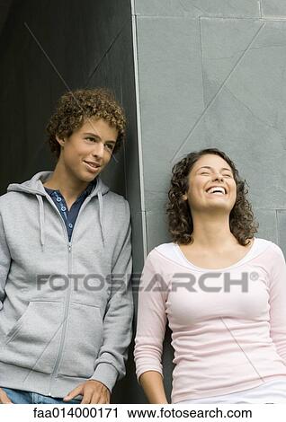 Stock Image - Young man and woman, leaning against corner of wall, laughing. Fotosearch