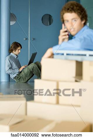 Stock Image - Young man sitting on floor using laptop, cardboard boxes in foreground. Fotosearch