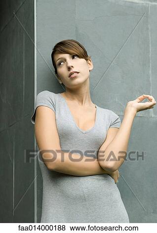 Stock Photo - Young woman leaning against wall, arm across body, looking away. Fotosearch