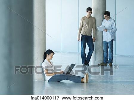 Stock Photo - Young woman sitting on floor using laptop, two men standing takling about document in background. Fotosearch