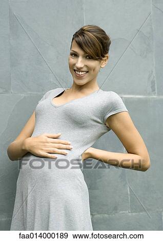 Stock Photo - Young woman standing, one hand on stomach and the other on back, smiling at camera. Fotosearch