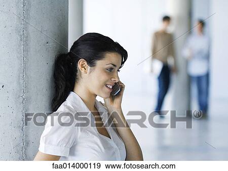 Stock Photo - Young woman using cell phone, colleagues standing in background. Fotosearch