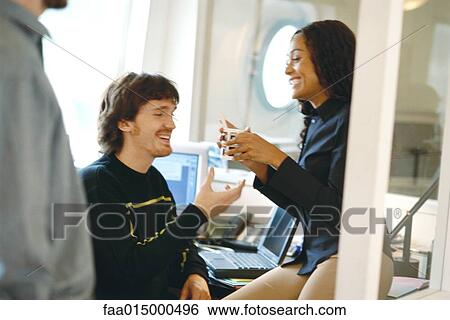 Colleagues in office, woman sitting on edge of desk handing man mug View Large Photo Image Stock Photograph - Colleagues in office, woman sitting on edge of desk handing man mug. Fotosearch