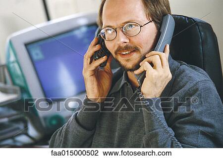 Stock Image - Man in office using cell phone and landline phone at same time. Fotosearch