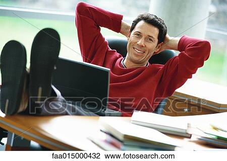 Man sitting at desk with feet up, holding laptop on lap, hands behind head View Large Photo Image Stock Image - Man sitting at desk with feet up, holding laptop on lap, hands behind head. Fotosearch
