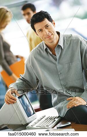 Man sitting on edge of desk, resting arm on computer, smiling at camera View Large Photo Image Stock Image - Man sitting on edge of desk, resting arm on computer, smiling at camera. Fotosearch