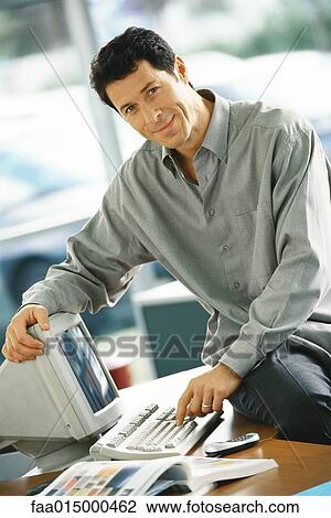 Man sitting on edge of desk, using computer, smiling at camera View Large Photo Image Stock Image - Man sitting on edge of desk, using computer, smiling at camera. Fotosearch