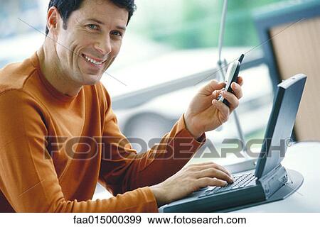 Man using laptop and dialing cell phone, smiling at camera View Large Photo Image Stock Photo - Man using laptop and dialing cell phone, smiling at camera. Fotosearch