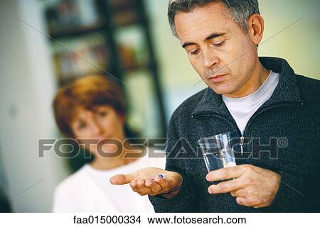 Senior man holding glass of water and pill, woman standing in background View Large Photo Image Picture - Senior man holding glass of water and pill, woman standing in background. Fotosearch
