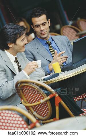 Two businessmen sitting at sidewalk cafe, using laptop View Large Photo Image Stock Image - Two businessmen sitting at sidewalk cafe, using laptop. Fotosearch