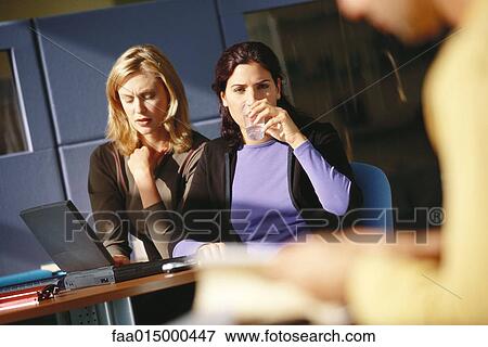 Stock Photo - Two female office workers using laptop, one drinking glass of water. Fotosearch