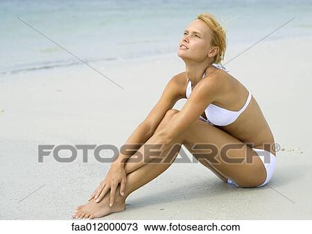 Stock Image - Woman in bikini sitting on beach, hugging knees, full length. Fotosearch