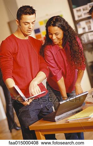 Young man and woman standing next to laptop, man pointing to user's manual View Large Photo Image Stock Photo - Young man and woman standing next to laptop, man pointing to user's manual. Fotosearch
