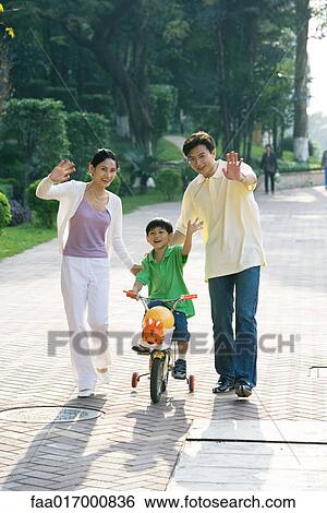 Stock Photograph - Boy riding bicycle, parents on either side, waving at camera. Fotosearch