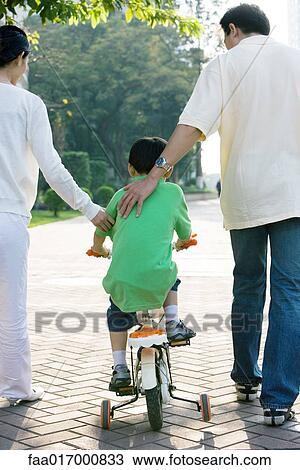 Stock Image - Boy riding bicycle with training wheels, parents on either side, rear view. Fotosearch