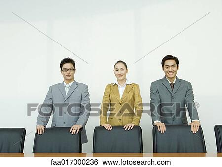Stock Photograph - Business executives standing with hands on backs of chairs in conference room. Fotosearch