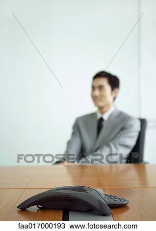 Businessman sitting at table, focus on conference phone in foreground View Large Photo Image Stock Image - Businessman sitting at table, focus on conference phone in foreground. Fotosearch