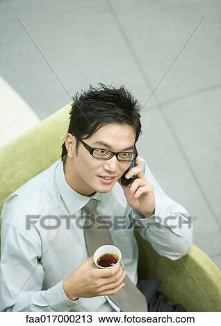 Stock Image - Businessman sitting in armchair, using cell phone and holding cup of coffee. Fotosearch