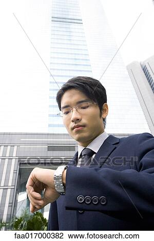 Stock Image - Businessman standing checking time on wristwatch, skyscraper in background. Fotosearch