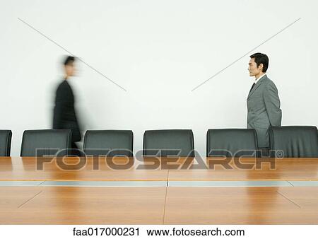 Stock Image - Businessman standing, facing associate arriving in conference room. Fotosearch