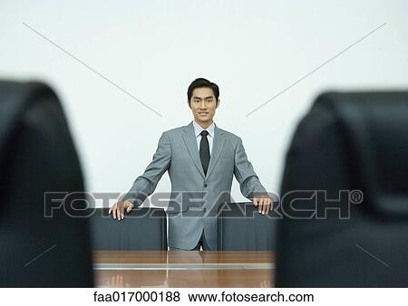 Stock Photo - Businessman standing in conference room with hands on backs of chairs, portrait. Fotosearch