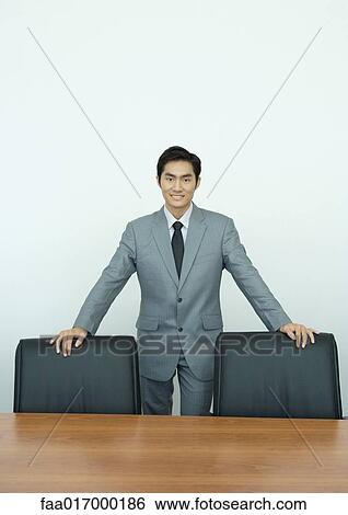 Stock Photograph - Businessman standing in conference room with hands on backs of chairs, portrait. Fotosearch