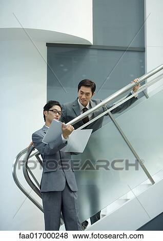 Businessman standing on stairs, looking down at file held by associate View Large Photo Image Stock Photo - Businessman standing on stairs, looking down at file held by associate. Fotosearch