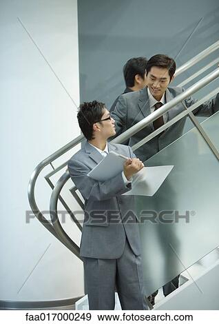 Stock Photo - Businessman standing on stairs, looking down at file held by associate. Fotosearch