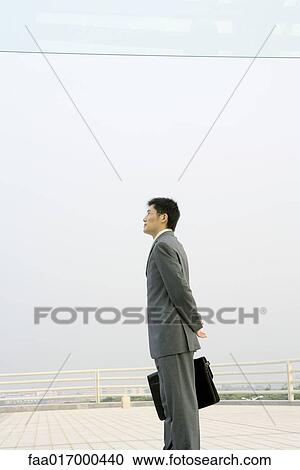 Stock Image - Businessman standing on terrace, looking toward distance. Fotosearch