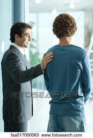 Stock Image - Businessman standing with hand on shoulder of casually dressed young man, smiling. Fotosearch
