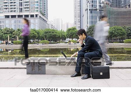 Picture - Businessman using laptop in office park, passers-by blurred in background. Fotosearch