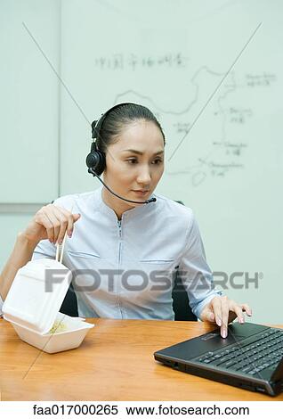 Stock Photography - Businesswoman eating takeout food, wearing headset, and using laptop. Fotosearch