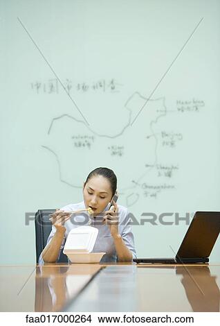 Picture - Businesswoman sitting at desk eating takeout food and using cell phone. Fotosearch