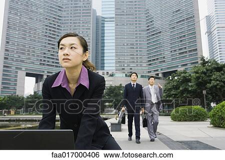 Stock Photograph - Businesswoman using laptop in office park, two businessmen approaching in background. Fotosearch