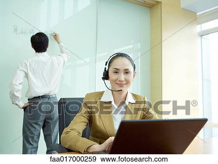 Businesswoman wearing headset and using laptop, colleague writing on white board in background View Large Photo Image Stock Photo - Businesswoman wearing headset and using laptop, colleague writing on white board in background. Fotosearch