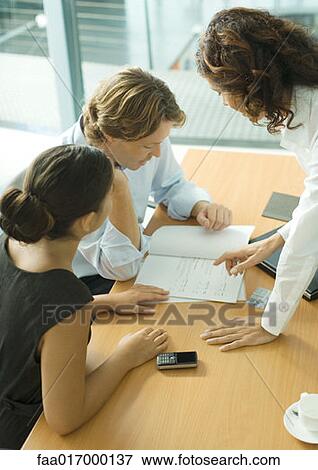 Stock Photo - Couple sitting at table looking at documents, professional woman standing across from them, pointing to page. Fotosearch