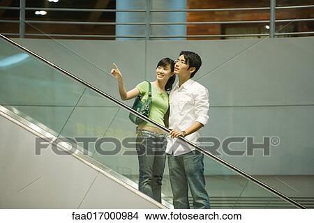 Couple taking escalator in shopping mall, woman pointing out of frame View Large Photo Image Picture - Couple taking escalator in shopping mall, woman pointing out of frame. Fotosearch