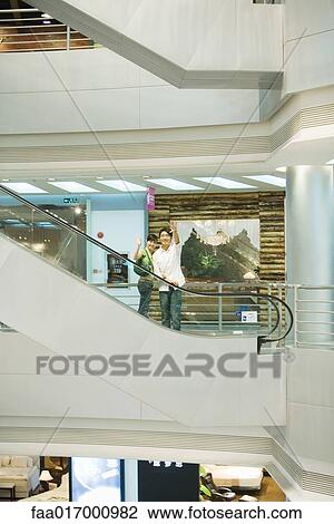 Stock Image - Couple taking escalator in shopping mall, waving at camera, in mid distance. Fotosearch