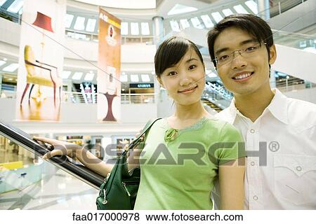 Stock Photo - Couple taking escalator in shopping mall, smiling at camera. Fotosearch