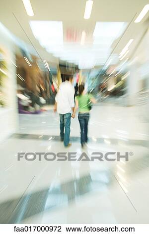 Stock Image - Couple walking in shopping mall, rear view, blurred motion. Fotosearch