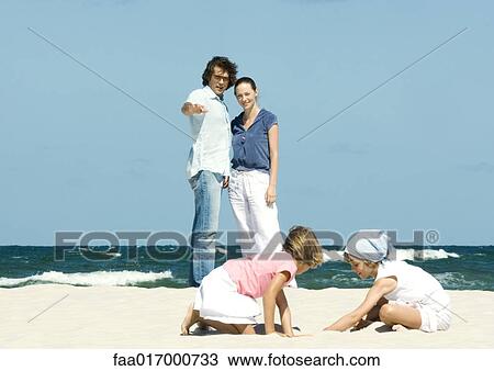 Stock Image - Family on beach, parents watching daughters play in sand. Fotosearch