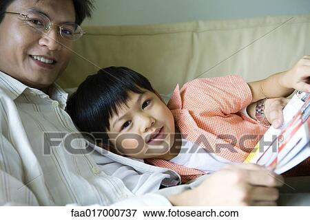 Stock Photo - Father and son sitting on sofa with book, smiling at camera. Fotosearch