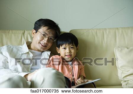 Stock Photography - Father and son sitting on sofa with book, smiling at camera. Fotosearch