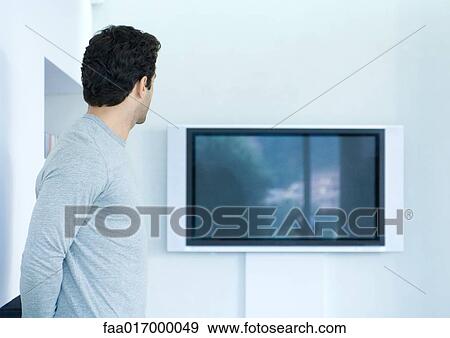 Stock Photo - Man looking toward flat screen TV on wall. Fotosearch