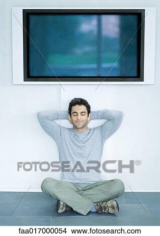 Man sitting on floor under wide screen TV, hands behind head and eyes closed View Large Photo Image Picture - Man sitting on floor under wide screen TV, hands behind head and eyes closed. Fotosearch