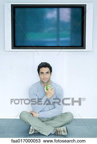 Stock Image - Man sitting on floor under wide screen TV, holding apple. Fotosearch