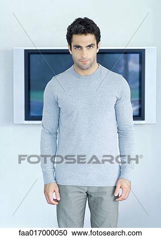 Stock Image - Man standing in front of wide screen TV, looking at camera. Fotosearch